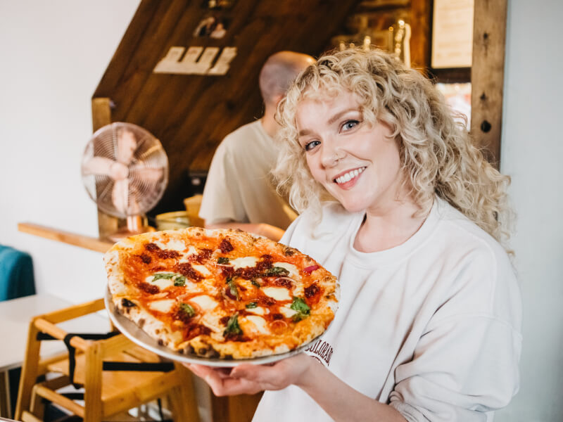A woman smiling holding handmade pizza up at a cooking class.